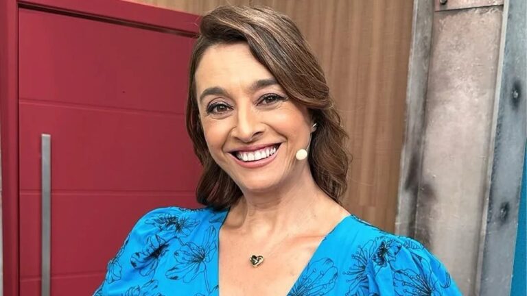 Mulher sorridente com microfone preso ao rosto veste um vestido azul com estampa floral preta e colar dourado em formato de coração, posando em um estúdio com parede de madeira e porta vermelha ao fundo.