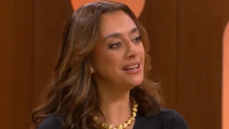 Mulher com colar dourado e blusa preta fala sorrindo durante gravação em estúdio de programa de TV