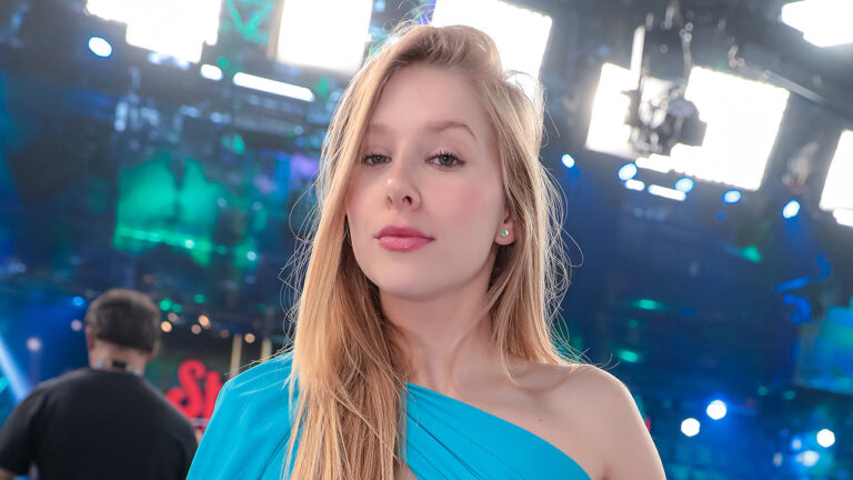 Influenciadora Cela Lopes de vestido azul ombro só posa no palco do Programa Silvio Santos com luzes de estúdio ao fundo e câmeras de televisão desfocadas