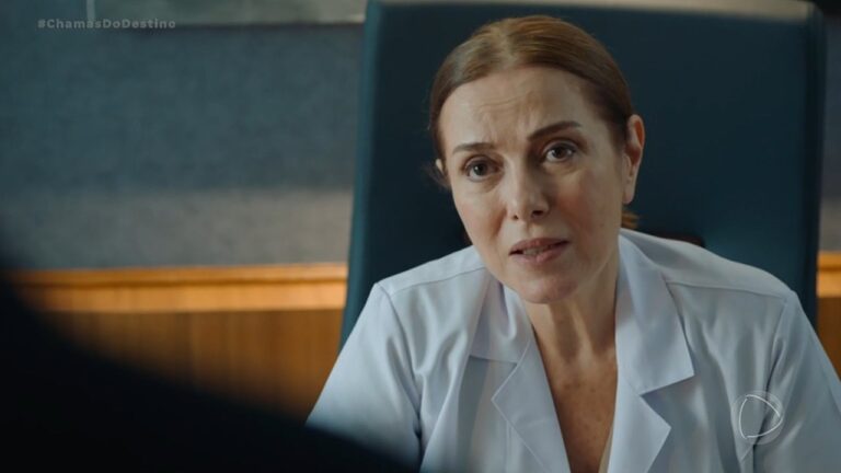 Mulher vestida com jaleco branco está sentada e conversa com expressão séria em ambiente que parece ser um consultório ou escritório. No canto superior esquerdo, aparece a tag da novela Chamas do Destino, exibida pela Record