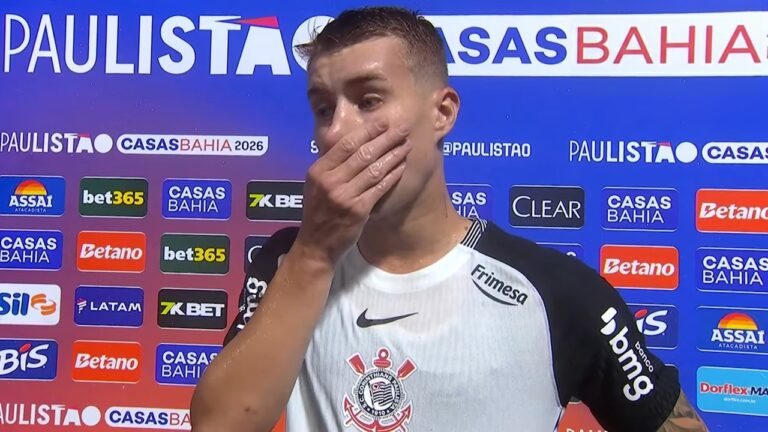 Charles, jogador do Corinthians, no intervalo da partida contra o RB Bragantino, concede entrevista na zona mista do Campeonato Paulista. Ele veste o uniforme do time e cobre parcialmente a boca com a mão, em frente a um painel com logotipos de patrocinadores como Casas Bahia, Betano e Assaí