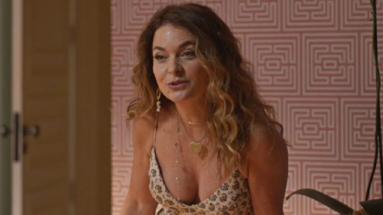 Mulher de cabelos castanhos cacheados e longos aparece inclinada levemente para a frente, usando vestido claro com estampa floral e colares dourados. Ela está em um ambiente interno com parede de padrão geométrico em rosa e branco ao fundo, aparentando estar conversando com alguém fora de quadro.