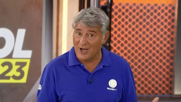 Homem de cabelos grisalhos usa camisa polo azul com logotipo da Record TV, falando em estúdio de programa esportivo com fundo colorido.