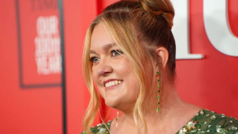 Mulher sorri em um evento com fundo vermelho e logotipos brancos parcialmente visíveis. Ela usa um vestido verde com bordados brilhantes, brincos longos de pedras verdes e tem o cabelo preso em um coque alto com algumas mechas soltas na frente.