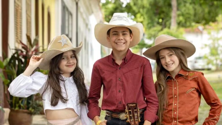 Três crianças sorridentes usando chapéus de caubói posam ao ar livre em uma rua arborizada com casas ao fundo. A menina à esquerda veste blusa branca de mangas bufantes e segura a aba do chapéu com uma das mãos, o menino ao centro usa camisa vermelha e cinto com fivela grande, segurando um violão, e a menina à direita veste camisa marrom com detalhes dourados, sorrindo para a câmera.
