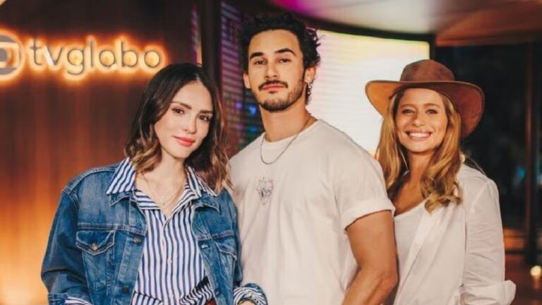 A imagem mostra três pessoas posando sorridentes em frente a um letreiro iluminado da TV Globo. Eles parecem estar em um evento ou lançamento, com trajes casuais e um clima descontraído.