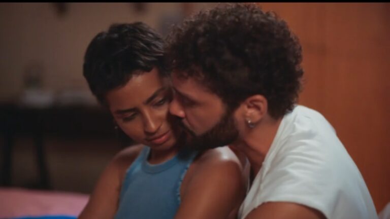 Homem beija o rosto de mulher sentada em cama enquanto ela baixa o olhar em cena íntima com iluminação quente em ambiente interno