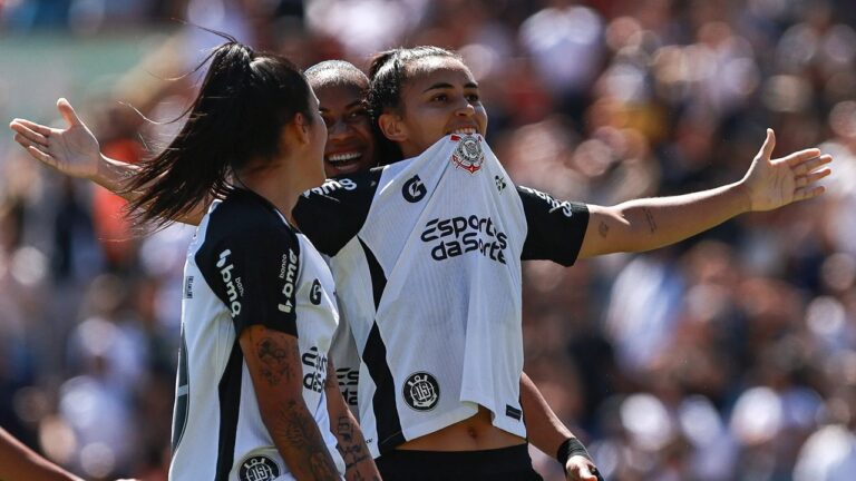 Três jogadoras de futebol comemoram em campo usando uniformes brancos e pretos com o escudo do Corinthians. Uma delas levanta a camisa para mostrar o símbolo do time enquanto sorri e abre os braços, sendo abraçada pelas companheiras. Ao fundo, há arquibancadas cheias de torcedores desfocados.