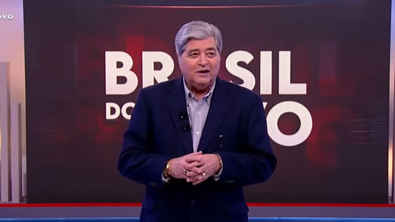 José Luiz Datena de terno azul escuro e mãos dadas no palco do programa Brasil do Povo da RedeTV! com o nome da atração ao fundo