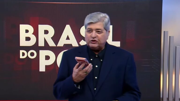 Homem de cabelos grisalhos fala segurando um celular em um estúdio de televisão. Ele veste paletó escuro e camisa preta. Atrás dele há um telão com o texto “BRASIL DO POVO” em letras grandes e brancas sobre um fundo vermelho e preto.
