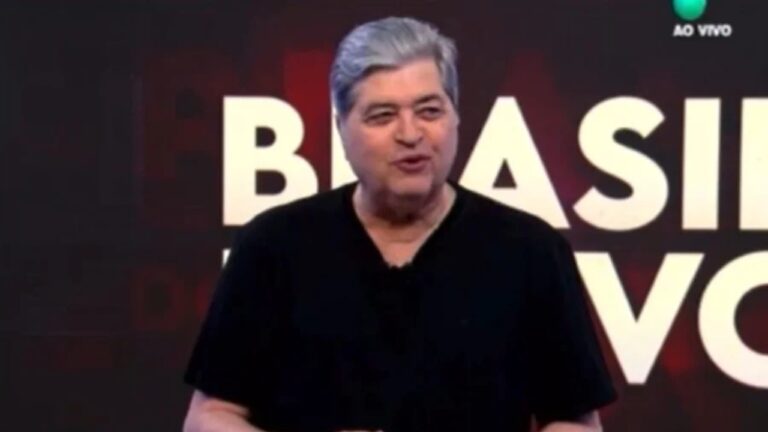 Texto Alternativo disse: Homem adulto em estúdio de televisão em plano médio com pele clara cabelos grisalhos curtos penteados para trás rosto arredondado expressão descontraída e levemente sorridente falando durante transmissão ao vivo vestindo camiseta preta de mangas curtas sem estampas com postura relaxada e braços próximos ao corpo ao fundo painel escuro em tons de preto e vermelho