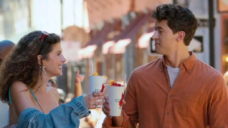 Mulher e homem jovens caminham em rua comercial ao ar livre durante o dia trocando sorrisos e brindando com copos grandes de bebida gelada decorados com frutas ela à esquerda tem cabelos castanhos longos e ondulados usa óculos apoiados na cabeça brincos longos top claro e jaqueta jeans caída nos ombros enquanto segura o copo com canudo e fatias de fruta