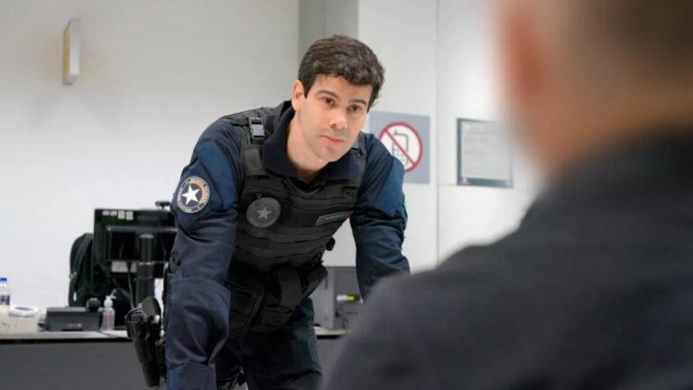 Homem com uniforme tático e colete à prova de balas inclina-se sobre uma mesa durante uma conversa séria em um ambiente de escritório, sugerindo uma cena de investigação policial.