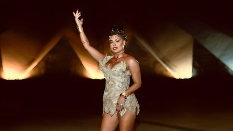 Deborah Caetano posa com vestido claro e acessórios dourados erguendo o braço em apresentação noturna com iluminação cênica ao fundo