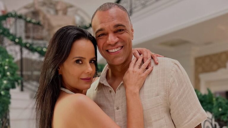 Casal posa abraçado e sorridente em um ambiente interno elegante, com decoração de escada e corrimão adornado por folhagens verdes ao fundo. A mulher usa um vestido claro e olha para a câmera, enquanto o homem, também de roupa clara, sorri amplamente.