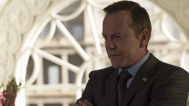 Homem de expressão séria veste terno escuro, camisa azul e gravata estampada, com um pequeno broche da bandeira dos Estados Unidos na lapela. Ele está de braços cruzados em um ambiente iluminado por luz natural, com janelas grandes e molduras brancas decorativas ao fundo.