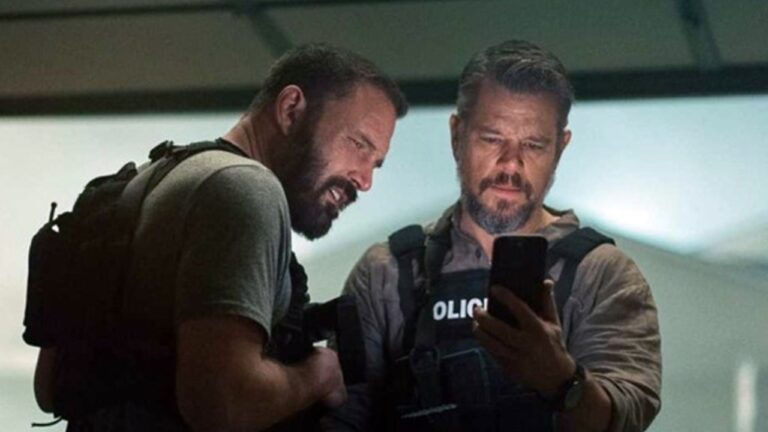 Dois homens com coletes táticos olham para um celular em ambiente fechado e com pouca luz