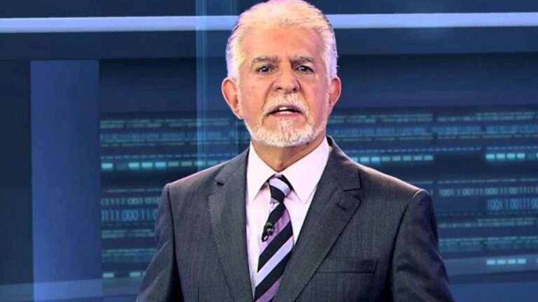 Homem mais velho, de cabelos e barba grisalhos, veste terno escuro com camisa branca e gravata listrada em tons de roxo e preto. Ele está em um estúdio de televisão, com um fundo azul repleto de elementos gráficos e linhas que remetem a um cenário de telejornal.