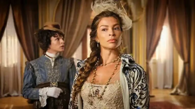 A imagem mostra duas pessoas vestidas com roupas de época em um cenário que lembra um palácio ou uma casa nobre. A mulher em primeiro plano usa um vestido rendado, colares longos e um penteado elaborado com uma trança lateral e um adorno de penas. Ao fundo, outra pessoa, também em trajes antigos, observa a cena, vestindo um casaco azul com rendas brancas e chapéu. O ambiente tem cortinas luxuosas e iluminação dourada, reforçando o clima histórico.