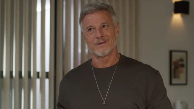 Homem de cabelos grisalhos curtos e barba aparada aparece sorrindo levemente em um ambiente interno bem iluminado. Ele usa uma camiseta escura e um colar prateado com pingente pequeno. Ao fundo, há cortinas verticais e uma parede com moldura decorativa.