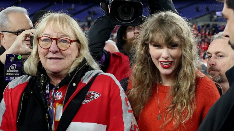 Duas mulheres sorridentes em um estádio lotado durante um evento esportivo da NFL, com câmeras e pessoas ao fundo. A mulher à esquerda usa óculos, casaco vermelho e preto com o emblema do Kansas City Chiefs e credencial pendurada no pescoço. A mulher à direita veste um suéter vermelho e batom combinando, com cabelos longos e ondulados.