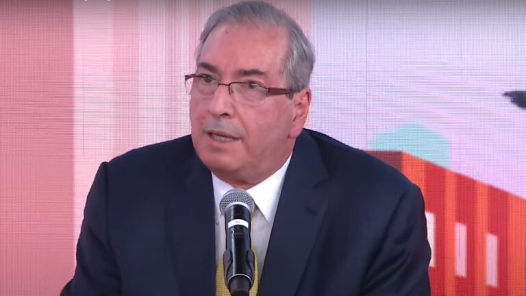 Homem de terno escuro e gravata amarela fala ao microfone durante evento ou entrevista, com fundo colorido em tons de vermelho e rosa.