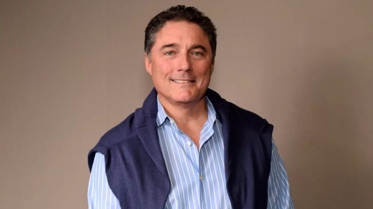 Homem de cabelos curtos e escuros sorri levemente enquanto posa diante de um fundo neutro. Ele usa camisa azul clara com listras brancas e um suéter azul marinho apoiado sobre os ombros, transmitindo aparência casual e elegante.