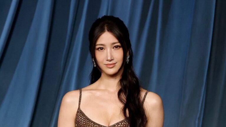 Mulher de cabelos longos e escuros posa diante de um fundo azul com tecido drapeado. Ela veste um vestido marrom brilhante com alças finas e decote em “V” e usa brincos prateados compridos, olhando para a câmera com expressão serena.