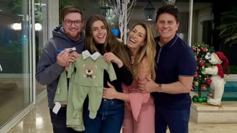 Grupo de quatro pessoas sorri enquanto segura roupinha de bebê e teste de gravidez em ambiente decorado com enfeites natalinos