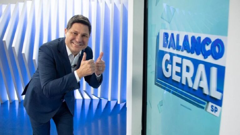 Apresentador do Balanço Geral SP sorri e faz sinal de positivo ao lado de telão com logotipo do programa em estúdio azul da Record