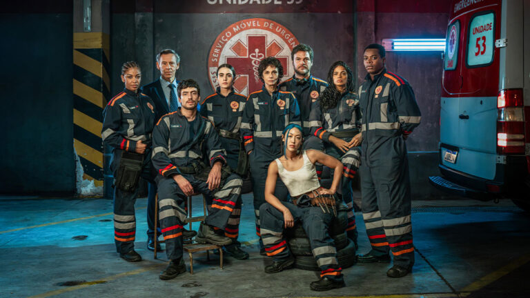 Atores de Emergência 53 posam sérios com uniformes de socorristas ao lado de ambulância no cenário da série médica da Globo