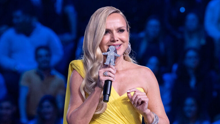 Eliana segura microfone com boneco do Criança Esperança durante apresentação em palco iluminado com plateia ao fundo em programa de auditório