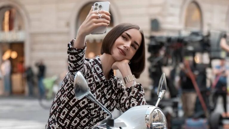 Mulher de cabelo curto e liso posa sentada em uma motocicleta branca enquanto tira uma selfie com o celular. Ela usa blusa estampada em preto e branco, bracelete prateado e expressão sorridente. Ao fundo, há prédios de arquitetura clássica e pessoas desfocadas, sugerindo uma rua movimentada de cidade europeia.