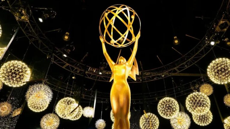 Grande estatueta dourada do Emmy, representando uma figura alada segurando uma esfera com anéis orbitais, iluminada sob luzes douradas em um palco decorado com esferas luminosas suspensas e estrutura metálica no teto.
