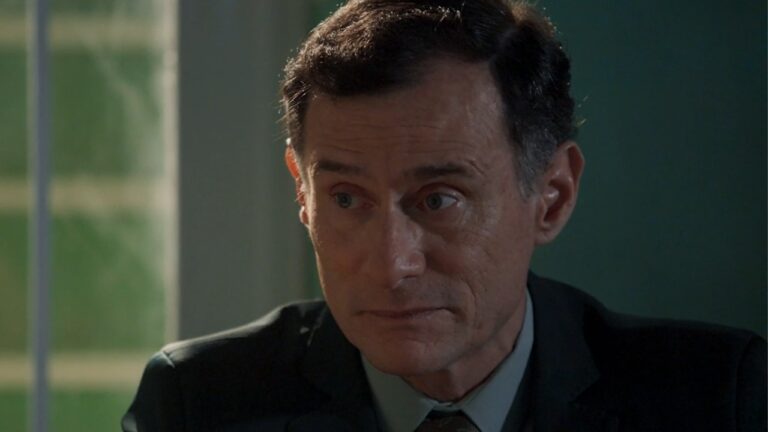 Homem de terno escuro olha para o lado com expressão séria em ambiente interno iluminado por luz natural