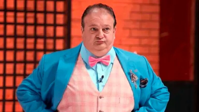 Homem com expressão séria veste terno azul, colete rosa xadrez e gravata borboleta rosa em estúdio de televisão