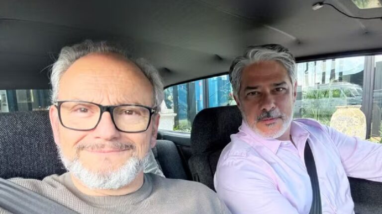 Dois homens estão sentados dentro de um carro, ambos usando cinto de segurança. O homem à esquerda, de óculos e barba grisalha, sorri levemente para a câmera, enquanto o homem à direita, também grisalho e de barba curta, veste camisa rosa clara e olha em direção à lente. A cena é iluminada pela luz natural do dia, com a parte externa visível pelas janelas do veículo.