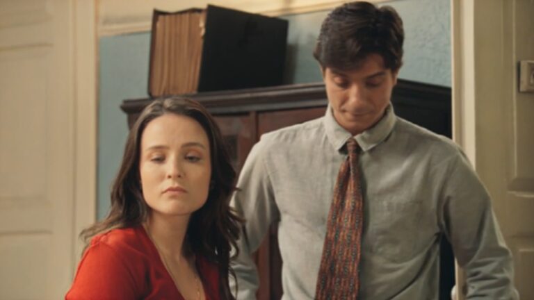 Mulher de cabelo castanho veste blusa vermelha e olha para o lado com expressão séria, enquanto homem ao seu lado, de camisa cinza e gravata colorida, mantém o olhar baixo com semblante preocupado. Eles estão em um ambiente interno com parede azul clara e móvel de madeira ao fundo, transmitindo um clima de tensão ou desconforto.