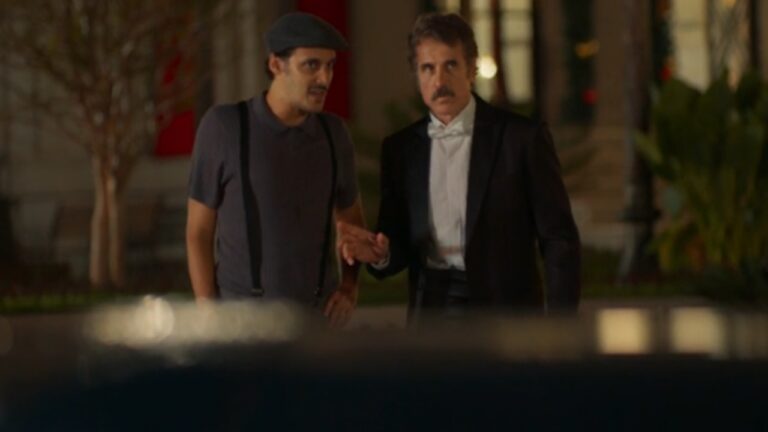 A imagem mostra dois homens conversando em um ambiente externo à noite. O homem à esquerda veste boina, camiseta escura e suspensórios, enquanto o da direita está de terno preto com camisa branca e gravata borboleta, aparentando estar mais formal. O fundo está desfocado, com um prédio iluminado e vegetação ao redor.