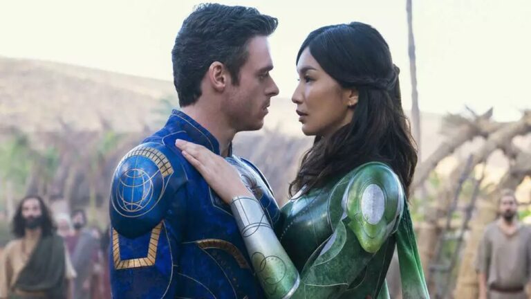 Cena de um filme de fantasia ou super-heróis mostrando um homem e uma mulher em trajes futuristas e coloridos — ele em azul e ela em verde — encarando-se de perto em um momento de intimidade. Ao fundo, há pessoas observando e uma paisagem árida com montanhas e vegetação seca.