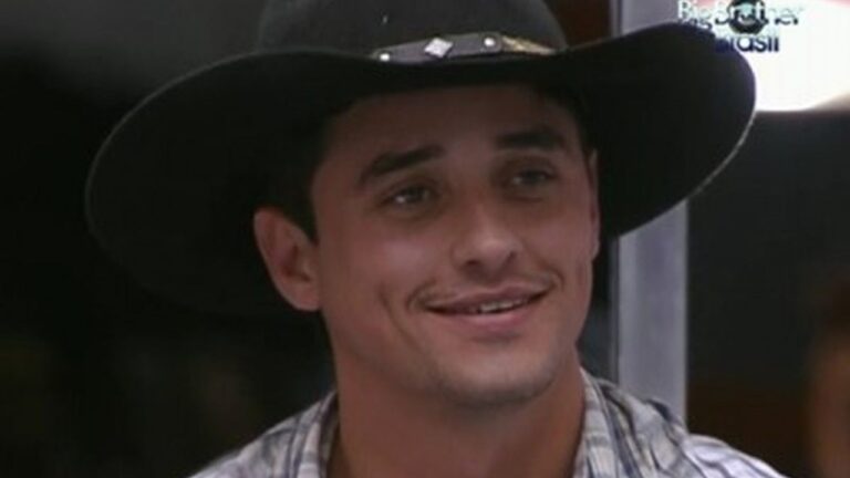 Homem sorri usando chapéu preto de aba larga e camisa xadrez em ambiente interno. No canto superior direito da imagem aparece o logotipo do programa Big Brother Brasil, indicando que a cena foi capturada durante uma edição do reality show.