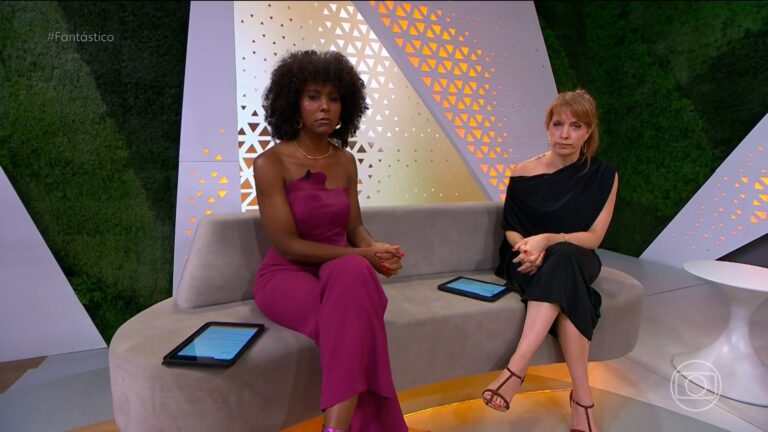 Maju Coutinho e Poliana Abritta, apresentadoras do programa Fantástico, da TV Globo, estão sentadas em um sofá de estúdio com design moderno e iluminação suave. Maju veste macacão roxo e Poliana, vestido preto de um ombro só; ambas têm tablets à frente e expressão séria durante o programa