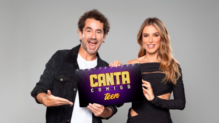 Felipe Andreoli e Rafa Brites posam sorridentes segurando um cartaz do programa Canta Comigo Teen, sobre um fundo neutro