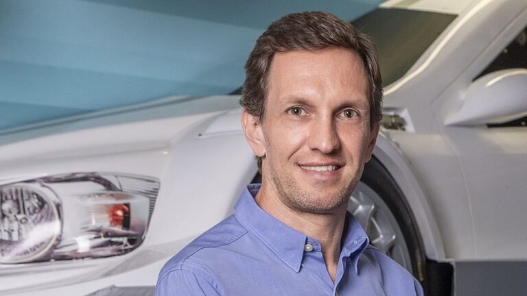 Homem sorri em ambiente automotivo, vestindo camisa azul clara; ao fundo, parte dianteira de um carro branco é visível sob iluminação suave.