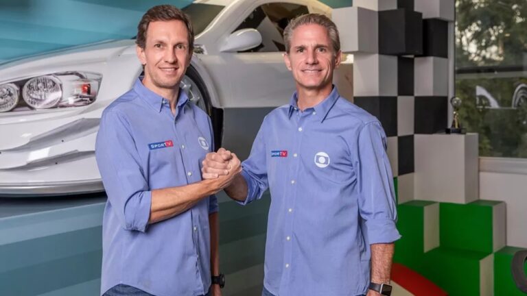 Dois homens posam sorrindo e apertando as mãos em estúdio decorado com temática automobilística, com parte dianteira de um carro branco ao fundo e elementos em formato de pódio nas cores preto, branco, verde e vermelho. Ambos vestem camisas azuis com logotipos da Rede Globo e do canal SporTV bordados.