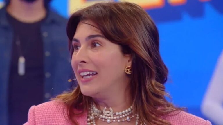 Mulher fala com expressão animada em um estúdio de televisão, usando microfone de lapela. Ela veste um conjunto rosa claro e colares de pérolas, além de brincos dourados. O fundo azul e colorido indica que está participando de um programa de TV.