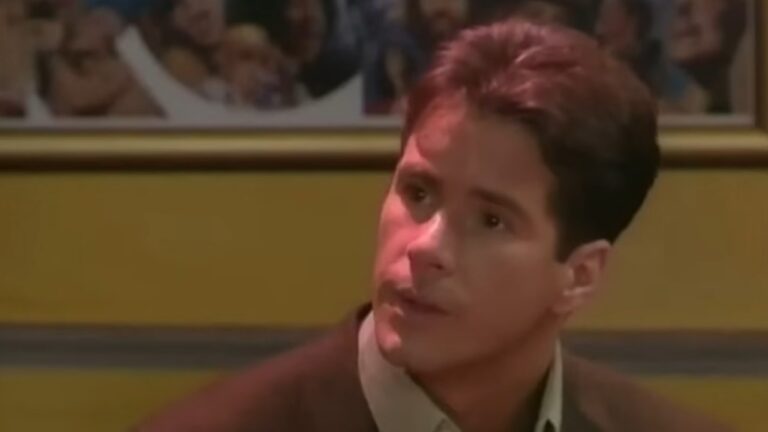 Homem com expressão séria olha para o lado em ambiente interno iluminado com luz amarelada. Ele veste camisa clara e casaco escuro. Ao fundo, há uma parede amarela com quadros contendo fotos coloridas emolduradas.