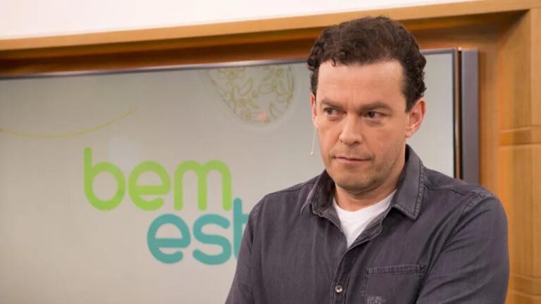 Homem aparece em um estúdio de TV, vestindo camisa cinza e camiseta branca por baixo, com um microfone de lapela preso ao rosto. Atrás dele há uma tela exibindo o logotipo do programa “Bem Estar”. Ele parece concentrado ou pensativo durante a gravação.