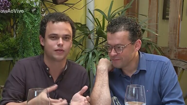 Dois homens sentados à mesa conversam em ambiente com decoração natural e taças à frente