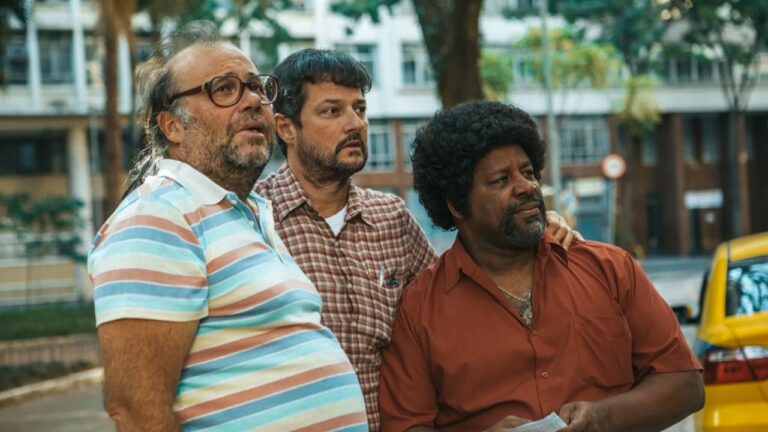 Três homens em rua arborizada observam algo fora de cena com expressão atenta enquanto prédio residencial aparece ao fundo